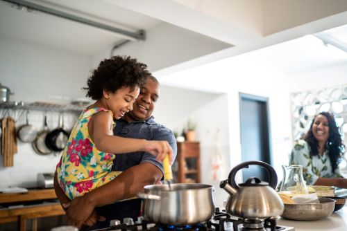 Girl in father's arms helping him cooking his new partner is watching them from the background: concept for How To Introduce Your Child To A New Partner