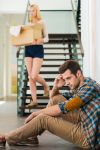 Frustrated young man sitting on the floor of his apartment while his girlfriend is leaving with boxes - BartonWood